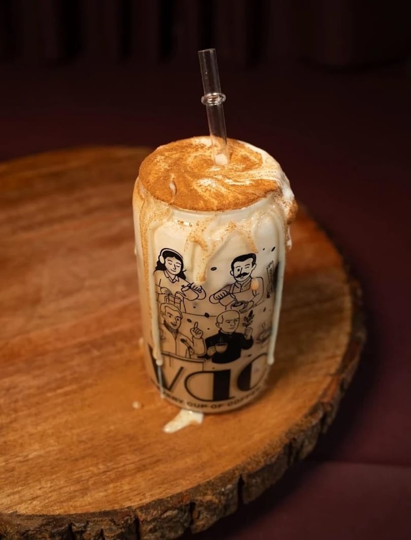 A tall glass of iced coffee with whipped cream, cinnamon, and a straw on a wooden slab.