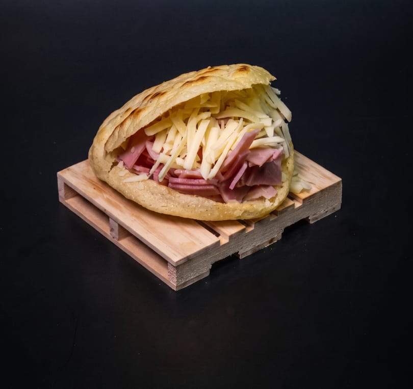 Arepa filled with shredded white cheese and sliced ham, served on a miniature wooden pallet.