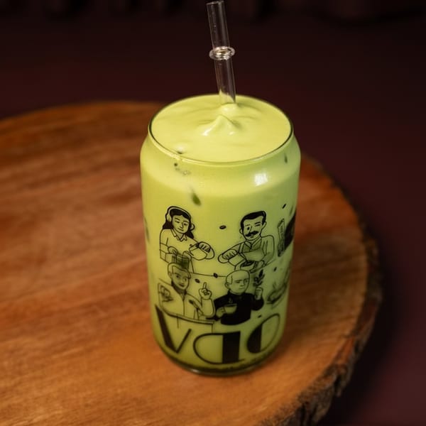 Green matcha latte with foam and a glass straw in a decorated glass on a wooden slab.