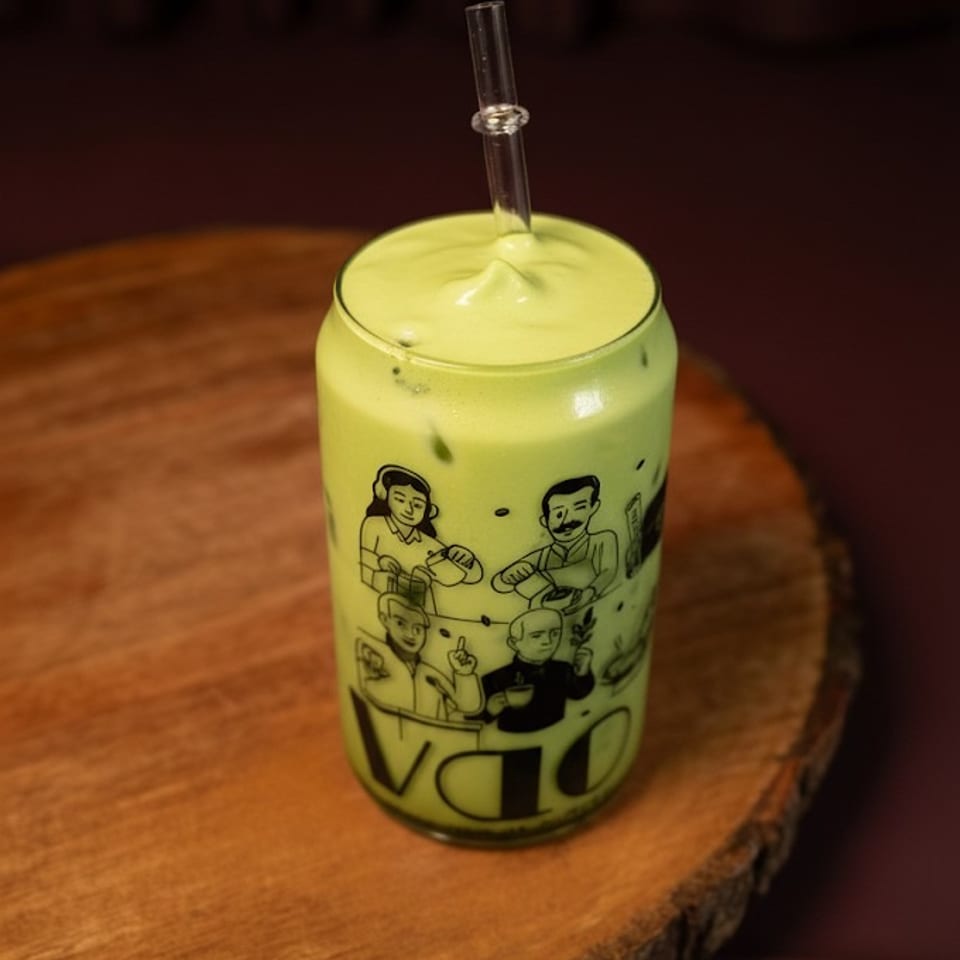 Green matcha latte with foam and a glass straw in a decorated glass on a wooden slab.