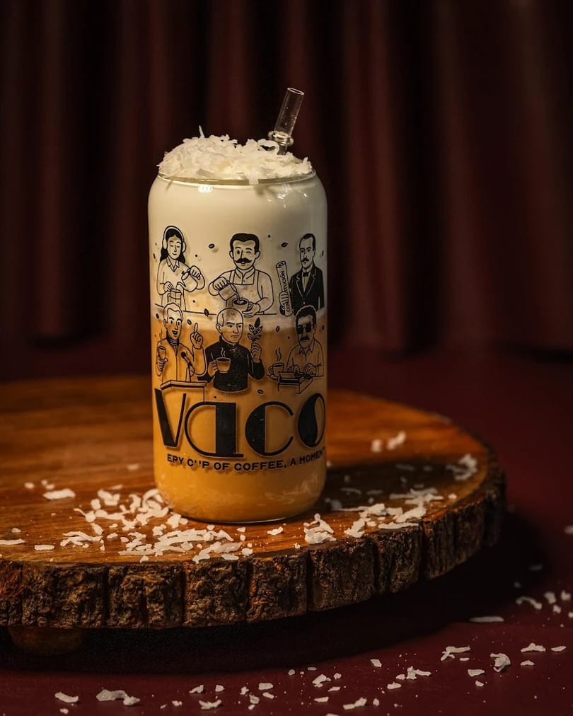 A Vaco branded glass with a layered drink, topped with whipped cream and coconut flakes.